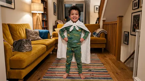 Photo 1: a small boy in dinosaur pajamas and a bedsheet cape, fists on his hips