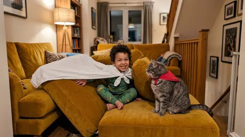 Photo 3: the boy peeking out of a den of sofa cushions beside a cat in a napkin cape