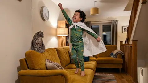 Photo 4: the boy standing on the sofa with one arm raised like a flying superhero