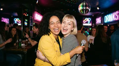 Photo 4: the two friends hugging and laughing under the neon lights